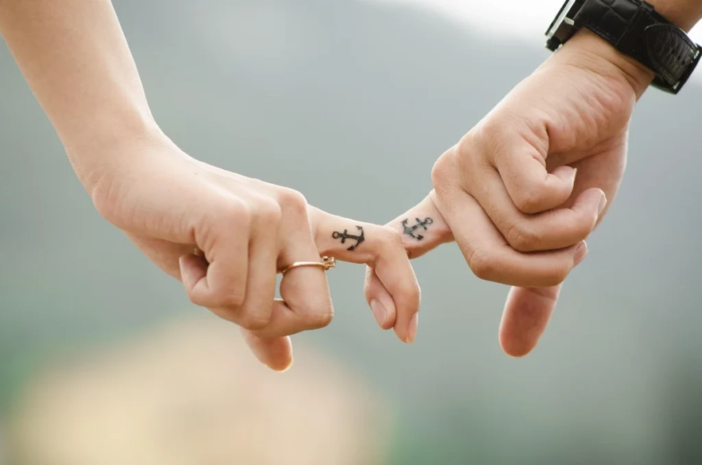 Two hands with anchor tattoos intertwined by pinky fingers, symbolizing connection and commitment.